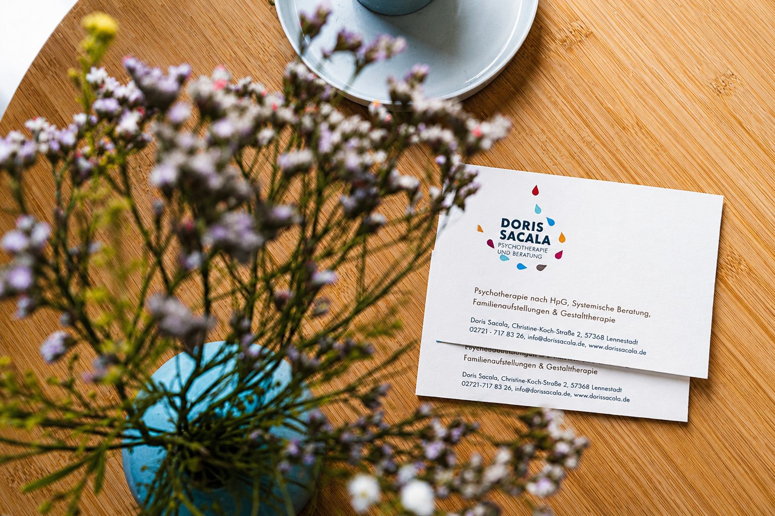 Visitenkarte der Praxis Doris Sacala auf einem Holztisch mit Blumen